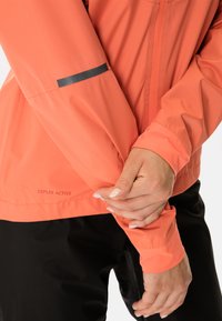 Korallfärgad vattentät jacka med slät textur, utrustad med dragkedja, svart detalj på ärmen och etiketten "CEPLEX ACTIVE" på fållen.