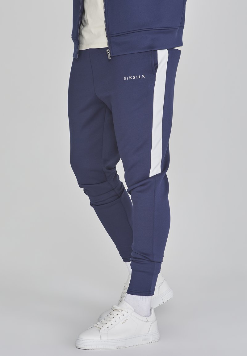 Jūras spēku sporta bikses ar baltām sānu svītrām, izgatavotas no mīksta auduma. Iezīmējas ar pieguļošu dizainu un SIKSILK logotipu priekšpusē. Nēsātas ar baltām sporta apaviem.