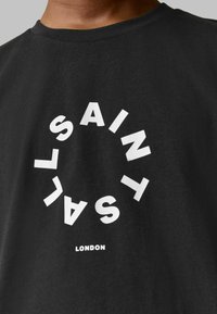Juodas medvilninis marškinėliai su ryškiu baltu tekstu, skaitančiu "SAINT" apskritoje formoje, o žemiau mažesnėmis raidėmis spausdinama "LONDON".