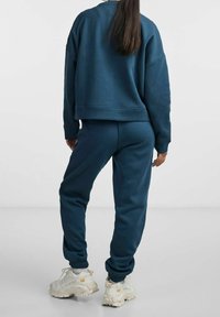 Person klädd i mörkblå sweatshirt och matchande sweatpants, vita kraftiga sneakers, står med ryggen mot betraktaren mot en enfärgad bakgrund.