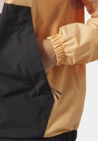 Chaqueta cortaviento con cuerpo naranja, mangas negras, puños elásticos y un bolsillo lateral con cierre de cordón. Textura suave.