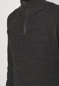 Nahaufnahme eines dunkelgrauen Strickpullovers mit gerippter Struktur und einem kurzen Reißverschluss am hohen Kragen.