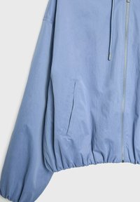 Veste zippée bleu clair avec ourlet élastique froncé, poche latérale et cordon suspendu à côté de la fermeture éclair sur un fond uni.