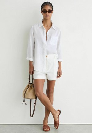 Kvinde iført en hvid skjorte med knapper, hvide shorts, solbriller, brune flettede sandaler, der holder en sandfarvet bucket bag og står op ad en ensfarvet væg.