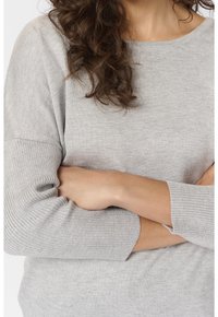 Personne aux cheveux bruns bouclés portant un pull gris clair à côtes, les bras croisés sur la poitrine.