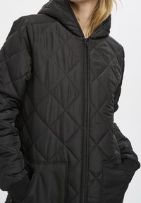 Chaqueta acolchada negra con capucha, cierre de cremallera y bolsillos laterales. El material es suave con un acabado mate y un patrón de diamantes.