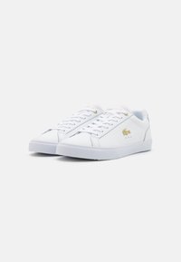 Vita sneakers i syntetiskt material, platta snören, gummisålar och en minimalistisk design. Har en guldlogotyp som accent och texturerade detaljer.