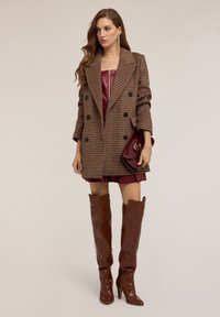 Abrigo de cuadros marrón de doble botonadura, vestido strapless de cuero burdeos y botas altas de cuero marrón con tacón, sosteniendo un clutch burdeos.