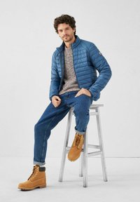 Giacca imbottita blu, maglione a motivo, jeans blu e stivali marroni con lacci. Modello seduto su uno sgabello di legno su sfondo chiaro.
