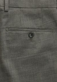 Gros plan d’un pantalon en tissu gris montrant une unique poche passepoilée arrière avec une fermeture à bouton noir.