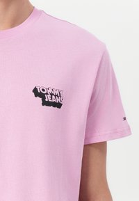 Rózsaszín pamut póló, melyen a bal mellen fekete "TOMMY JEANS" logó található. Rövid ujjú és kerek nyakú, minimális varrásokkal.