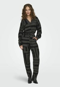 Zwarte jumpsuit met een geometrisch gouden lijnpatroon, lange mouwen en een trekkoord in de taille, gecombineerd met puntige zwarte enkellaarsjes.