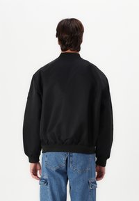 Giubbotto bomber nero con texture liscia, polsini e vita a costine. Presenta un colletto classico ed è abbinato a pantaloni cargo blu.