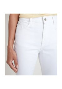 Close-up of a person wearing white denim jeans and a beige top, showing waist, button, pockets, and left hand resting by side.