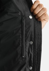 Schwarze gesteppte Jacke mit einem Reißverschlussfach, Druckknöpfen und einer glatten Stoffoberfläche. Das Innere ist sichtbar und zeigt das Futter.