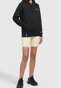 Czarna bluza z kapturem i żółtym logo, z kieszenią z przodu i teksturowanym materiałem; połączona z żółtymi szortami rowerowymi i czarnymi butami sportowymi.