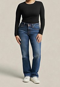 Femme portant un haut noir cintré à manches longues, un jean bleu coupe droite et des baskets blanches, debout devant un fond uni.