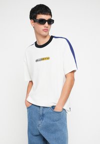 Tricou alb cu accente negre și albastre, având un logo galben și negru pe piept. Croială lejeră și mâneci scurte, material din bumbac.