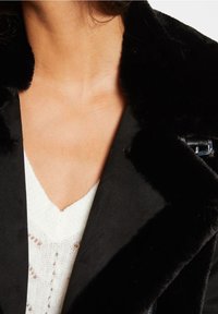 Černé, sametové vnější oblečení s měkkou texturou, pod ním je hluboký výstřih v bílé barvě se vzorovaným pleteným designem.