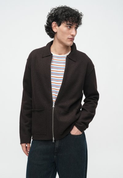 Brązowa kurtka zapinana na suwak, wykonana z miękkiego, teksturowanego materiału, z kołnierzem i dwoma przednimi kieszeniami. Nosi się ją na wzorzystą koszulę w odcieniach niebieskiego i pomarańczowego.