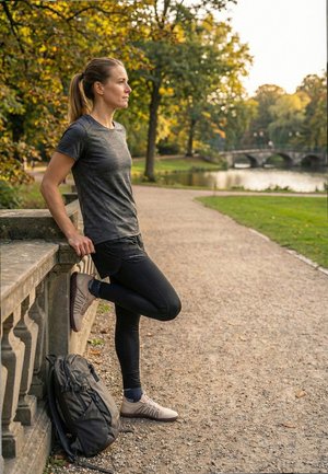 Mujer con ropa deportiva estirando la pierna contra una barandilla de piedra en un camino del parque, con una mochila en el suelo y árboles junto a un río en otoño.