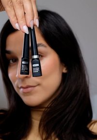 Deux tubes d'eyeliner noir au design élégant et rectangulaire, étiquetés "Eyeliner Brillant" et "Eyeliner Coloré," tenus devant un arrière-plan neutre.