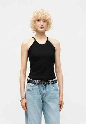 Jeune personne aux cheveux blonds bouclés portant un haut dos-nu noir, un jean bleu clair, une ceinture noire cloutée et un bracelet argenté.