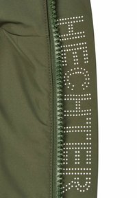 Olivgrüne Jacke mit einem Reißverschluss an der Seite und gepunkteter Schrift. Das Material wirkt glatt mit mattem Finish und minimalistischem Design.