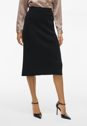 Jupe crayon en tricot noir à taille haute, avec un motif texturé. Associée à des escarpins noirs à bout pointu et un chemisier en satin marron clair.