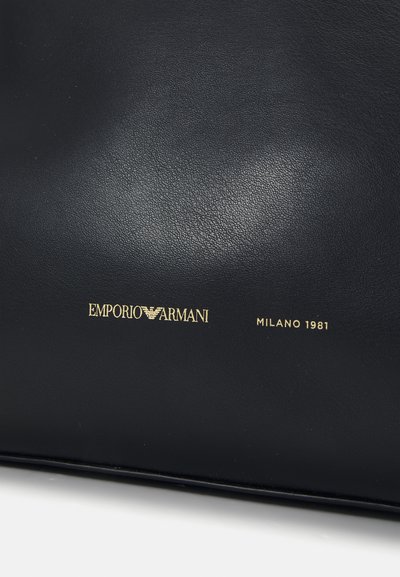 Melna ādas soma ar zelta reljefu logo "EMPORIO ARMANI" un "MILANO 1981" detaļu, gluda tekstūra, taisnstūra forma.