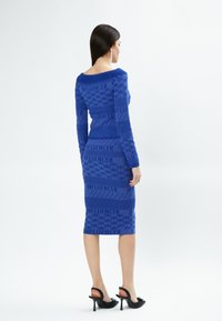 Abito in maglia blu con maniche lunghe, scollatura off-shoulder, pattern testurizzati e la scritta "INFLUENCER" ripetuta; lunghezza al ginocchio con silhouette aderente.