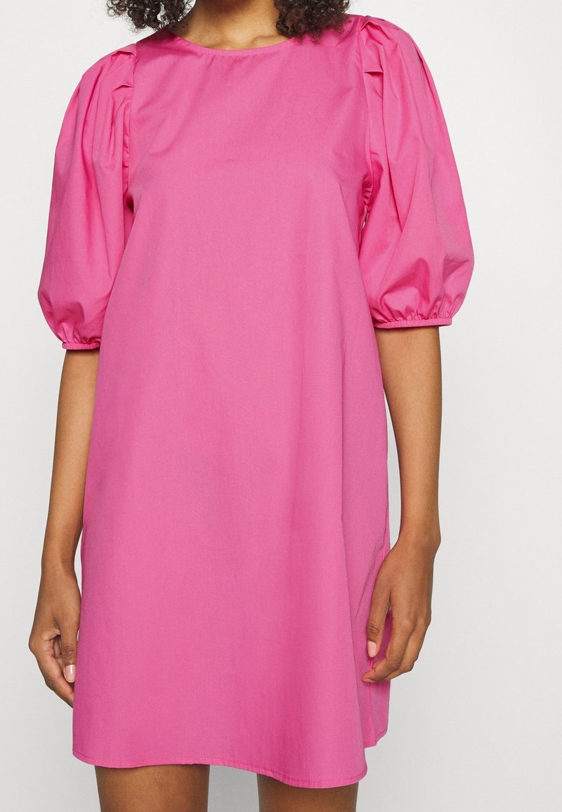 Femme portant une robe rose vif avec des manches bouffantes jusqu'au coude, debout les bras détendus devant un fond clair.