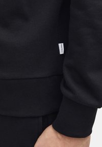 Black sweatshirt featuring ribbed cuffs and hem, made of textured fabric with a small brand label sewn into the side.
