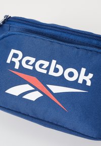 Bolsa azul marinho feita de tecido texturizado, apresentando um logotipo branco da Reebok e detalhes em vermelho; inclui um fecho zip na parte superior.