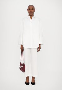 Camicia bianca oversize con maniche larghe, indossata con pantaloni bianchi lunghi. Gli accessori includono una piccola borsa bordeaux e scarpe nere con punta.