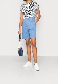 Donna con una maglietta a maniche corte a fantasia, pantaloni corti azzurri a vita alta, scarpe da ginnastica bianche, che tiene in mano una piccola borsa blu navy, in piedi contro uno sfondo semplice.