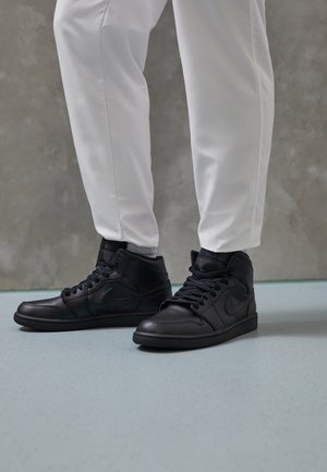 Personne portant un pantalon blanc et des baskets montantes noires se tenant sur un sol bleu clair contre un mur gris.