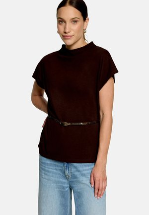 Jeune femme portant un haut marron foncé à manches courtes avec une fine ceinture et un jean bleu clair, debout devant un fond clair uni.