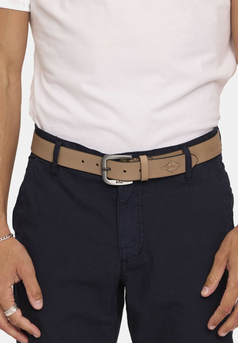 Tan leren riem met een zilveren metalen gesp, met een gestructureerd oppervlak en een logo-label. G gedragen bij een marineblauwe broek en een wit shirt.