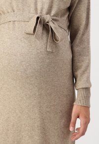 Vestito in maglia beige con vita legata, polsini a coste e una texture morbida. Il tessuto presenta un'apparenza leggermente melangiata.