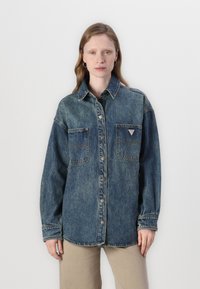 Denimskjorte i falmet blå med knappefront, to brystlommer og et logo-patch. Har lange ermer og klassisk krage.