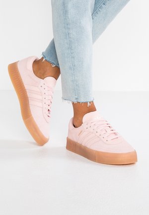 Osoba nosząca jasnoniebieskie dżinsy i różowe sneakersy na platformie z gumowymi podeszwami, stojąca na białej podłodze.