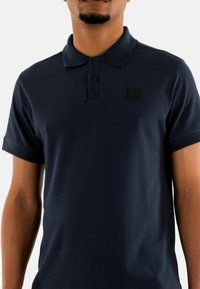 Marinblå pikétröja i strukturväv med treknappslås, korta ärmar och en liten svart logotyppatch på bröstet.