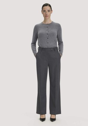 Frau trägt eine graue, geknöpfte Strickjacke, dunkle weite Hose und schwarze High Heels, steht mit Händen in den Taschen vor einem schlichten hellen Hintergrund.
