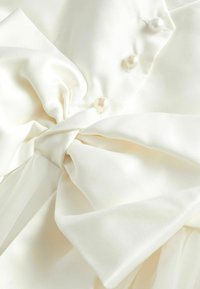 Tissu en satin blanc mettant en avant un grand nœud, orné de trois boutons en perles. Texture douce avec des sections superposées et flottantes.
