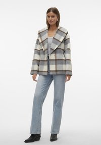 Cappotto a quadri con ampi rever in misto lana crema, grigio e marrone; chiusura con un bottone; abbinato a jeans azzurri chiari e stivali neri.