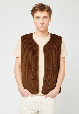 Gilet in corduroy marrone con texture a coste, scollo rotondo e chiusura con bottoni a pressione. Indossato sopra una t-shirt beige chiara.