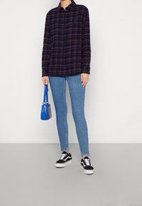 Camicia in flanella a quadri blu navy, maniche lunghe, con bottoni, abbinata a jeans skinny blu chiaro e sneakers nere. Tiene in mano una piccola borsa blu.