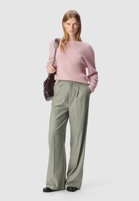 Różowy, puszysty sweter zestawiony z jasnoszarymi spodniami w prążki. Modelka trzyma ciemnobrązową torbę, ma na sobie czarne buty.