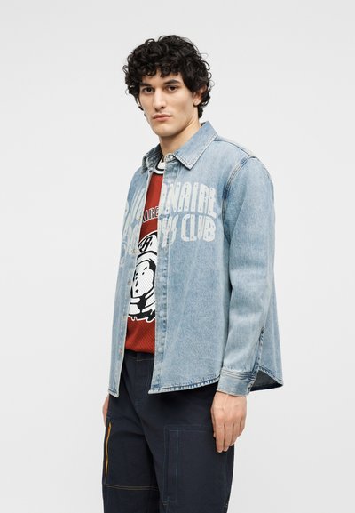 Άνδρας με σγουρά μαύρα μαλλιά, ντυμένος με μπλε τζιν μπουφάν πάνω από κόκκινη μπλούζα με λευκό κείμενο και σκούρο παντελόνι, κοιτάζει προς τα εμπρός με λευκό φόντο.